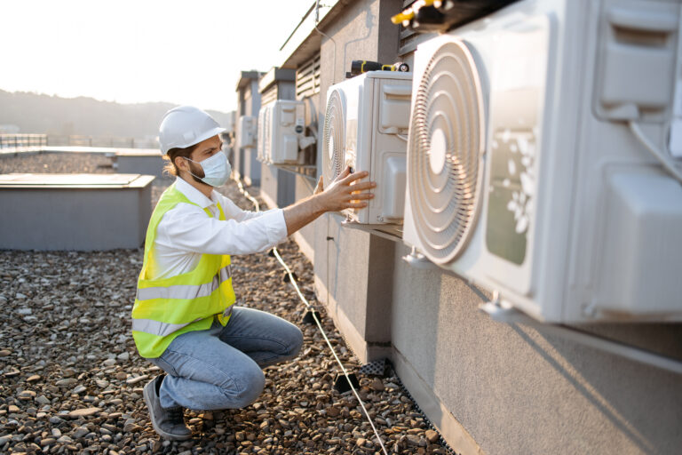 Professional worker of plant wearing dust mask crouching opposite air conditioner and providing inspection on fresh air. Caucasian bearded man checking attachment of device to wall in urban roof.