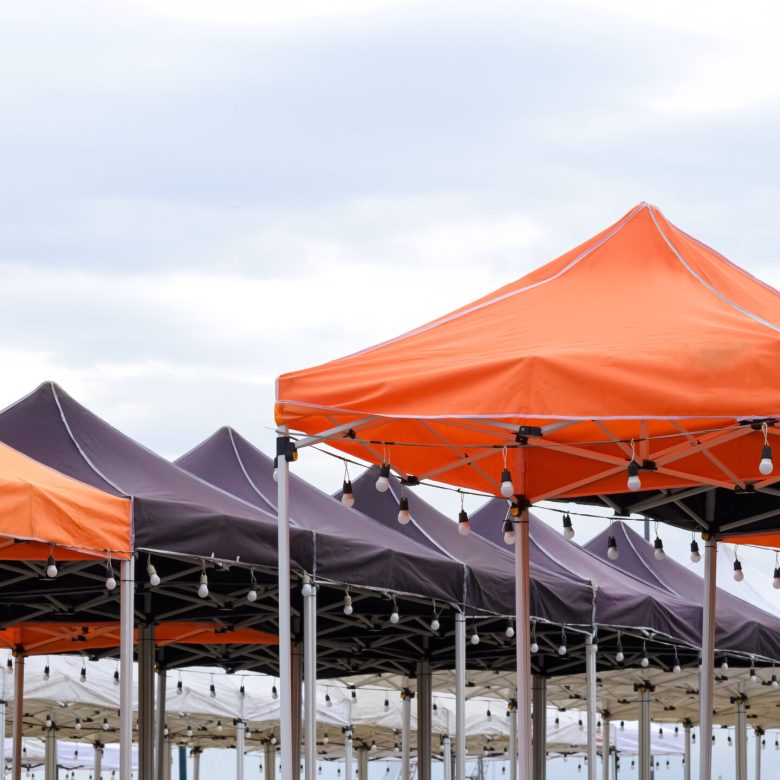 Rows of many foldable canopy tents with hanging LED light bulbs decoration for outdoors event against cloudy sky background